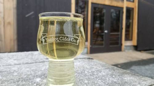 A glass of cider from Yankee Cider Co. sits on a stone surface, with a rustic building in the background.