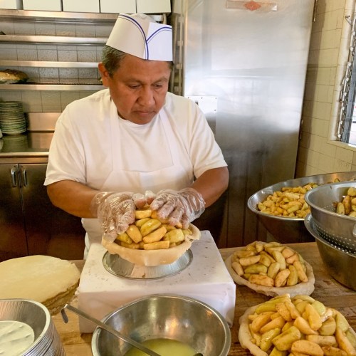 A chef in a white uniform and hat prepares a pie filled with bananas in a kitchen, surrounded by bowls and pastry shells.