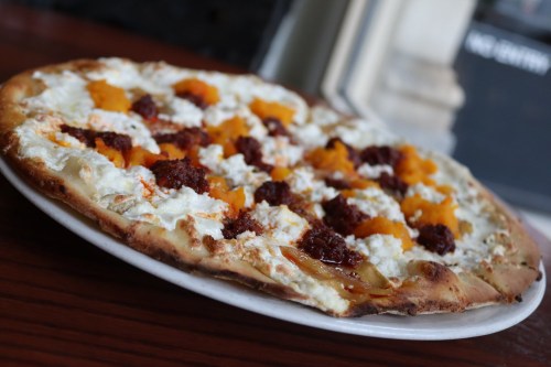 A close-up of a pizza topped with ricotta, chorizo, and orange squash on a wooden table.