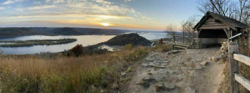 A panoramic view of a river valley at sunset, with a rustic shelter and grassy hillside in the foreground.