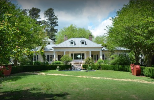 A large, elegant house with a metal roof, surrounded by lush greenery and a small fountain in the front yard.