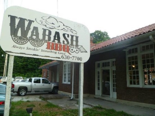 Sign for Warash BBQ, featuring the slogan "Always Smokin' Something Good," outside a brick building with parked cars.