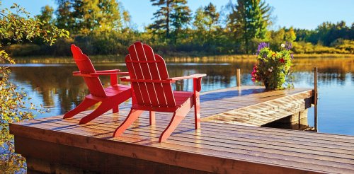 Two red Adirondack chairs on a wooden dock overlooking a calm lake, surrounded by trees and flowers in sunlight.