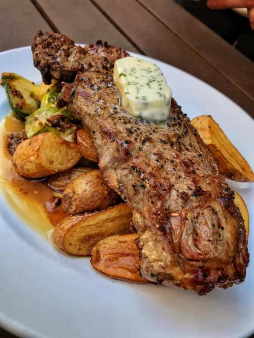 A juicy steak topped with herb butter, served with roasted potatoes and Brussels sprouts on a white plate.