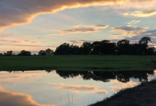 A serene landscape at sunset, featuring a calm pond reflecting colorful clouds and trees along the horizon.