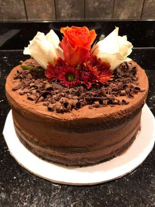 A chocolate cake topped with chocolate shavings and decorated with white and orange roses and red flowers.