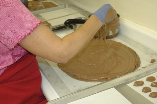 A person in a pink shirt and gloves spreads melted chocolate on a marble surface in a kitchen setting.