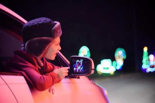 A child leans out of a car window, smiling at colorful holiday lights in the background at night.