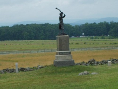 A soldier statue holding a rifle stands on a stone pedestal in a grassy field with distant hills.