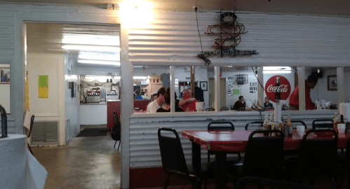 Interior of a casual restaurant with red tables, patrons dining, and a Coca-Cola sign on the wall.