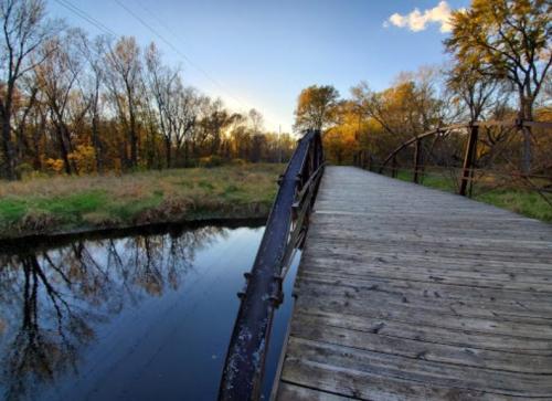 A wooden bridge over a calm river, surrounded by trees with autumn foliage under a clear blue sky.