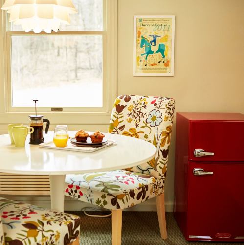 A cozy dining area with a round table, floral chairs, a red fridge, and a window overlooking a serene outdoor view.