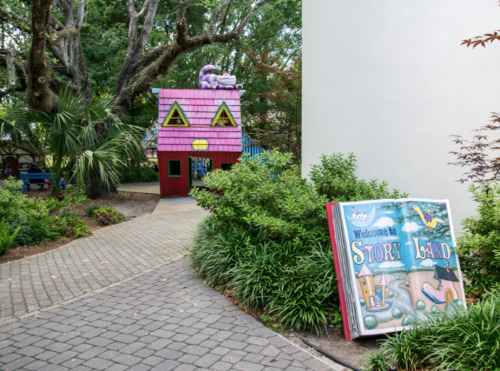 Colorful playhouse with a purple roof and a sign reading "Welcome to Story Land" in a lush garden setting.