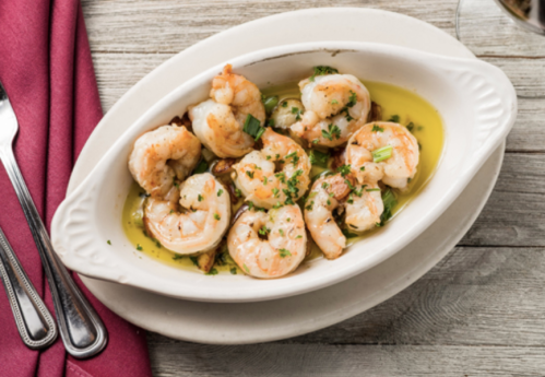 A dish of sautéed shrimp in olive oil, garnished with herbs, served in a white oval bowl on a wooden table.