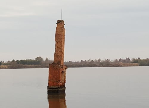 A weathered brick pillar stands in calm water, surrounded by trees and a cloudy sky.