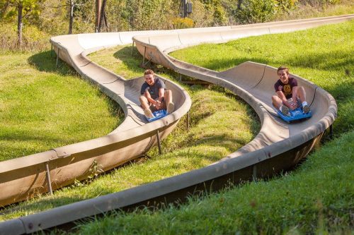 Two people enjoy a summer ride on a winding slide through green grass and trees.