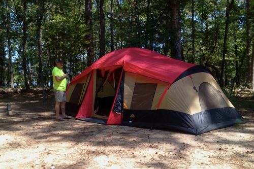 A person in a bright shirt stands next to a large red and tan tent in a wooded campsite.