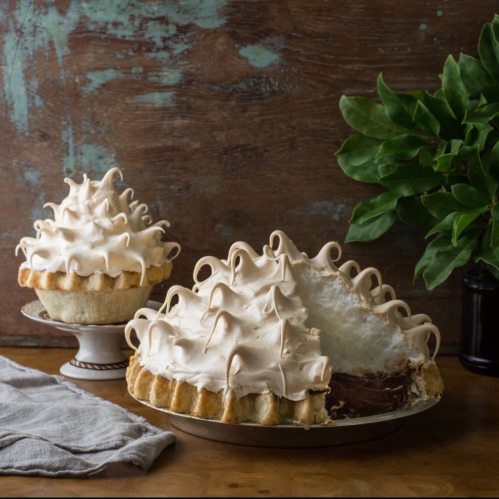 Two pies with fluffy meringue topping, one partially sliced, on a wooden table with a green plant in the background.