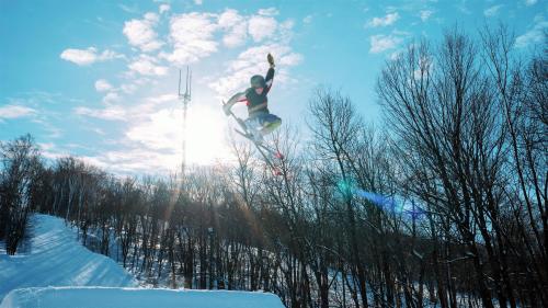 A skier performs a jump against a bright blue sky and snowy landscape, with trees and a tower in the background.