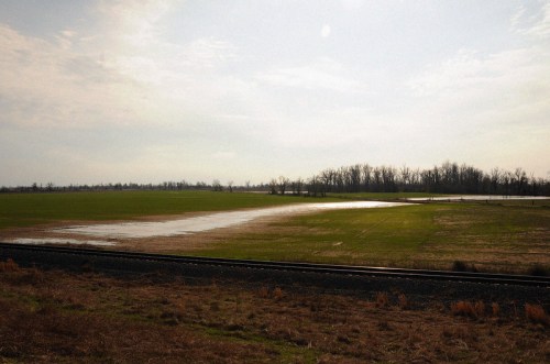 A wide, open field with patches of water, bordered by a railway track and distant trees under a cloudy sky.