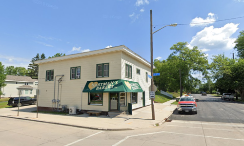 A corner bakery with a green awning, surrounded by trees and parked cars on a sunny day.