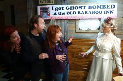 A group of four people reacts in surprise to a woman in a vintage dress, standing by a sign about being "ghost bombed."
