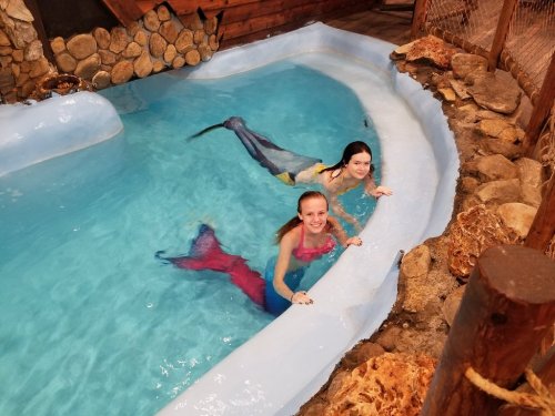Two girls in colorful mermaid tails swim in a blue pool surrounded by rocks and wooden decor.