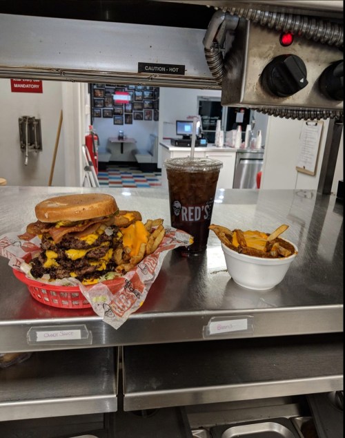 A large cheeseburger with bacon, fries in a bowl, and a drink on a counter at a diner.