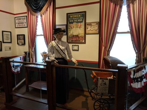 A museum display featuring a mannequin in a suffragette outfit, with historical posters and a baby carriage nearby.