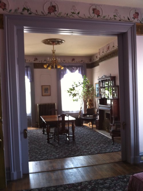 A view through a doorway into a vintage room with a table, chairs, and large windows, adorned with floral decor.