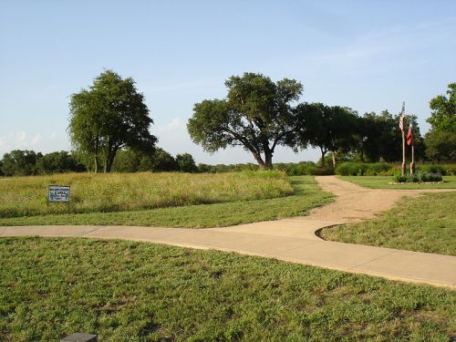 A grassy area with a winding path, trees, and a flagpole in the background under a clear blue sky.