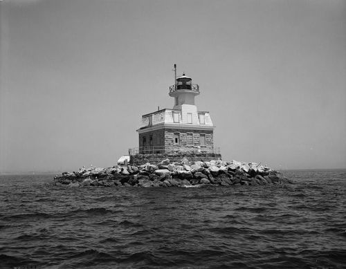 A historic lighthouse stands on a rocky island, surrounded by water, under a clear sky. Black and white photo.