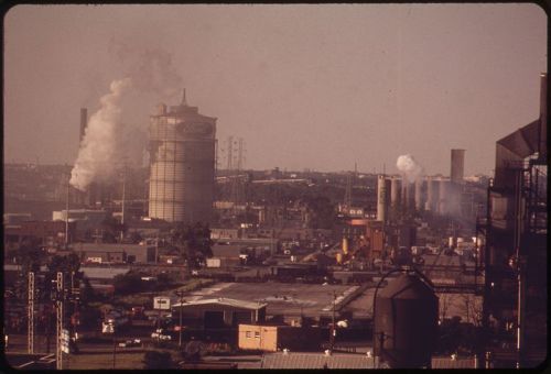 Industrial landscape with smokestacks and factories, emitting smoke against a hazy sky.