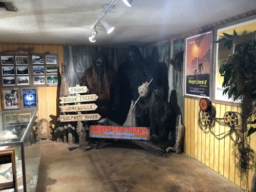 Display featuring a Bigfoot exhibit with signs pointing to local areas, posters, and a rustic interior in a museum setting.
