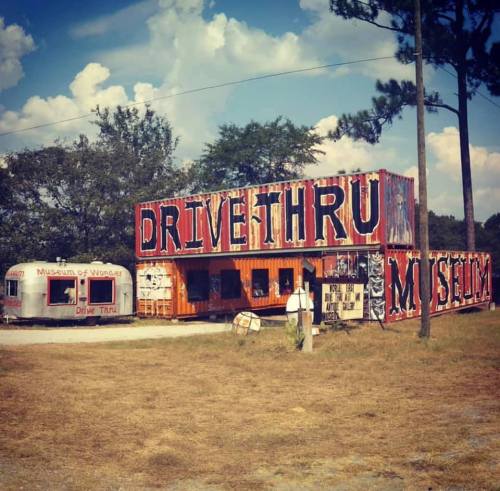 A colorful drive-thru museum with shipping containers and a vintage trailer, set against a blue sky and grassy area.