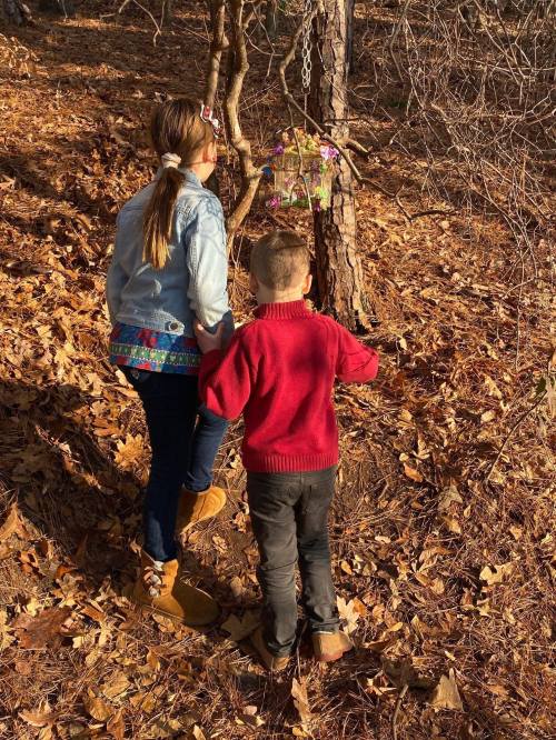 Two children explore a wooded area, looking at a colorful hanging decoration on a tree amidst fallen leaves.