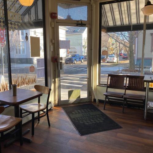A cozy café interior with wooden tables, a bench, and large windows showing a street view outside.