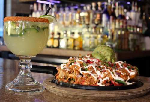 A vibrant margarita beside a sizzling platter of nachos topped with guacamole, sour cream, and fresh ingredients.