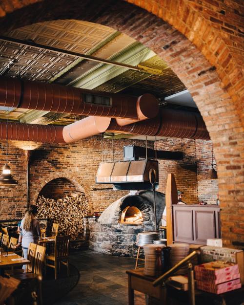 Cozy restaurant interior featuring a brick oven, wooden tables, and exposed ductwork in a warm, inviting atmosphere.