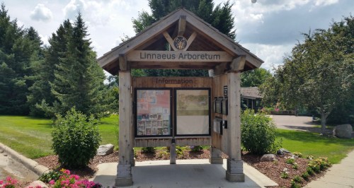 Information kiosk at Linnaeus Arboretum, surrounded by greenery and flowers under a partly cloudy sky.