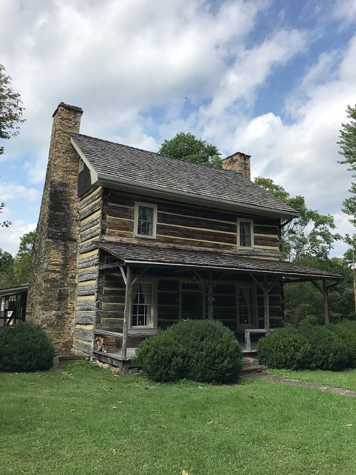 A rustic log cabin with a stone chimney, surrounded by greenery and under a partly cloudy sky.