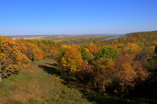 A scenic view of a river valley surrounded by colorful autumn trees under a clear blue sky.