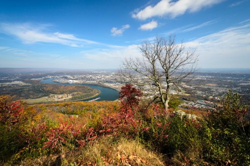 A scenic view from a mountain overlooking a river and a city, surrounded by autumn foliage and a clear blue sky.