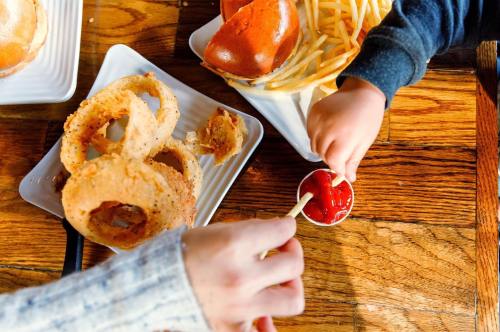 A child's hand reaches for ketchup while a plate of onion rings and fries sits on a wooden table.