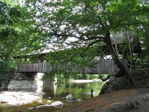 A wooden covered bridge spans a calm river, surrounded by lush green trees and rocky banks.