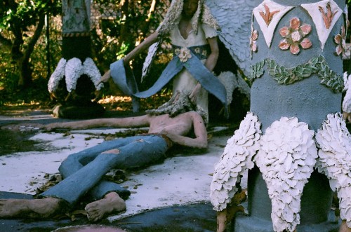 A surreal sculpture scene featuring a figure lying on the ground, surrounded by ornate, colorful decorations and foliage.
