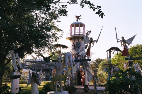 A whimsical outdoor sculpture garden featuring a lighthouse and various artistic figures with swords and birds.