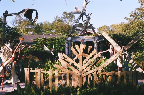 A whimsical outdoor scene featuring sculptures, wooden structures, and greenery under a clear blue sky.