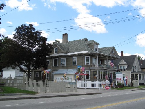 A large, two-story house with a gray roof, surrounded by a white picket fence and colorful flags, on a sunny day.