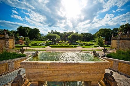 A sunlit garden with lush greenery, fountains, and a stone basin in the foreground, under a partly cloudy sky.